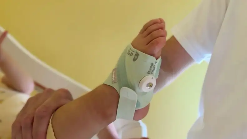 Baby's foot being held up in mint green Owlet Dream Sock in front of a yellow wall.