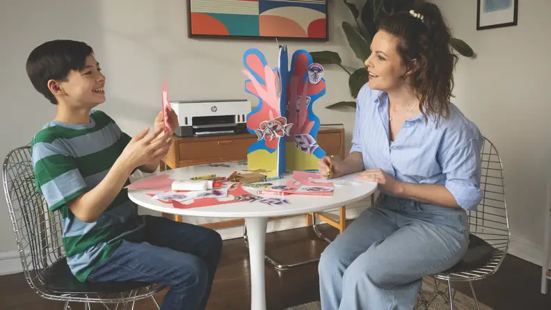 A mom and child doing arts and crafts at a table with an HP printer in the background.