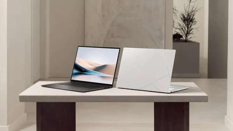 Two Asus Zenbook S 16 laptops, one with its screen facing the camera and one facing away, sitting on a table in a modern-styled room.