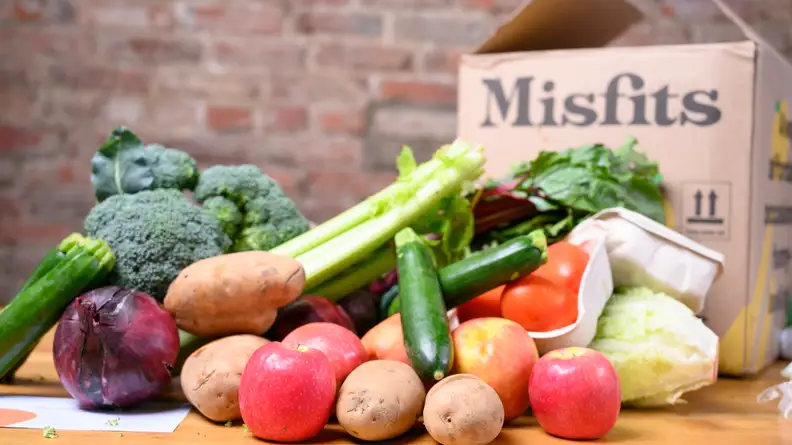 A pile of fresh produce such as potatoes, tomatoes, zucchini, celery, broccoli, red onion, lettuce, and apples sits in front of a Misfits Market box against a brick wall background.