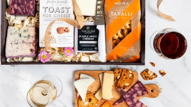 Food packaging with cutting board with cheese and crackers on top. Wine on the side.