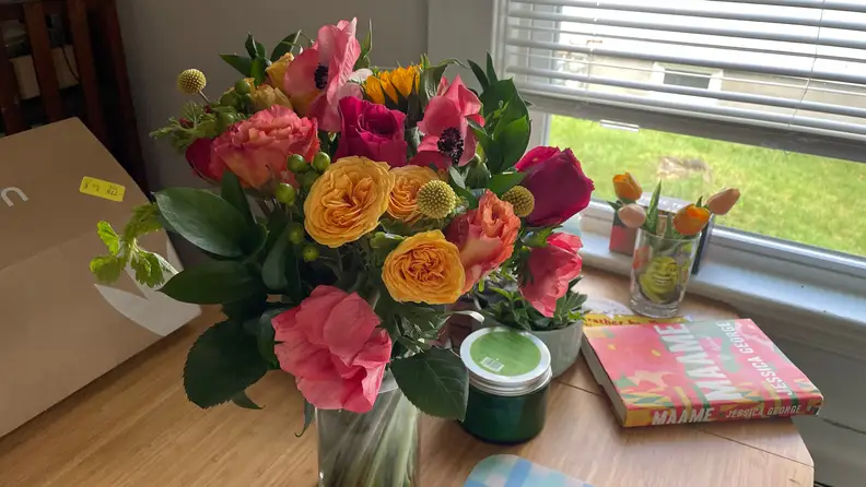 An UrbanStems bouquet on a wood table with a smaller vase of flower in the background, next to a window and book.