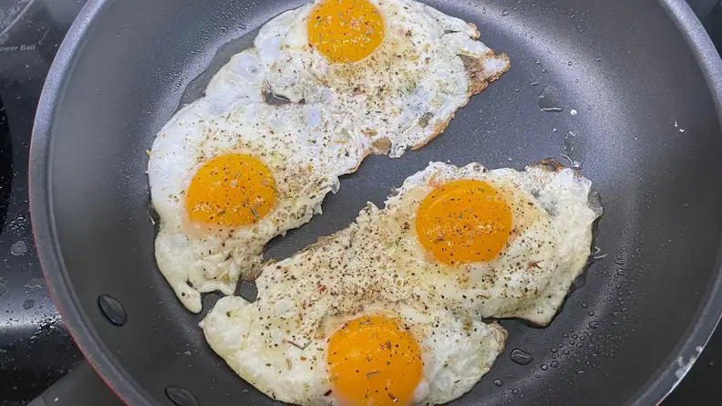 Sunny side up eggs frying inside of greasy skillet on cooktop.