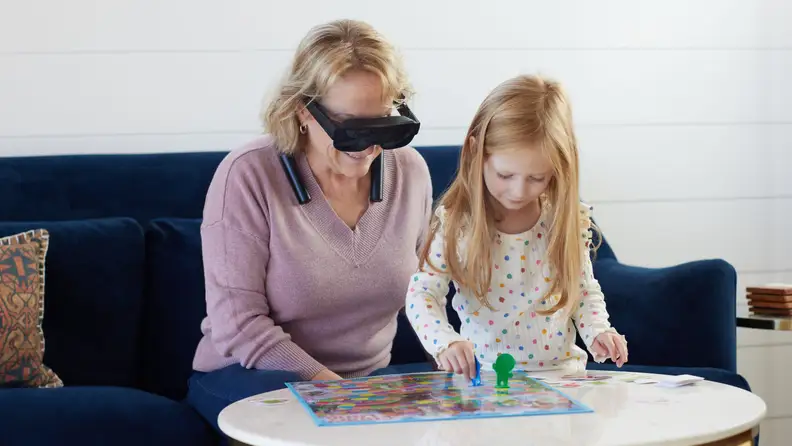 A grandmother sits on a couch while wearing eSight Go eyewear and watching a child play a board game.