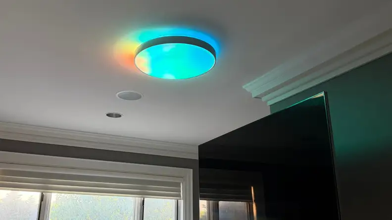 Flush-mount smart ceiling light displaying colorful ambient glow effects around the fixture, casting blue, green, and warm accent lighting onto the ceiling.