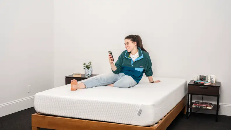 A woman sits on the Tuft & Needle Mattress on her phone