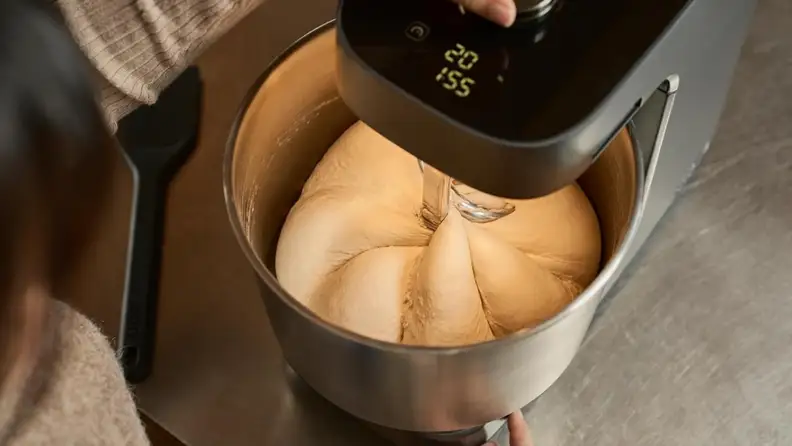 Top-down view of a stand mixer kneading bread dough in a stainless steel mixing bowl.