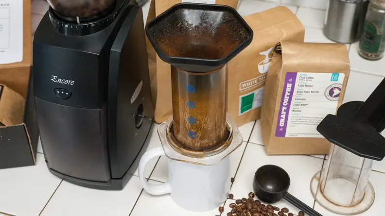 Assorted coffee gadgets arranged on a white surface, including a coffee mug, an Aeropress coffee maker, a coffee bean grinder, a measuring spoon, and coffee bean bags.