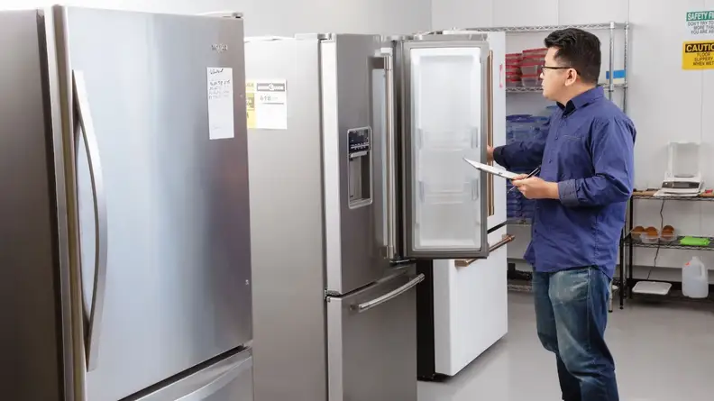 A man stands in a fridge testing lab with a clipboard in hand, in front of a row of refrigerators. He's holding one of their doors open, examining the interior.