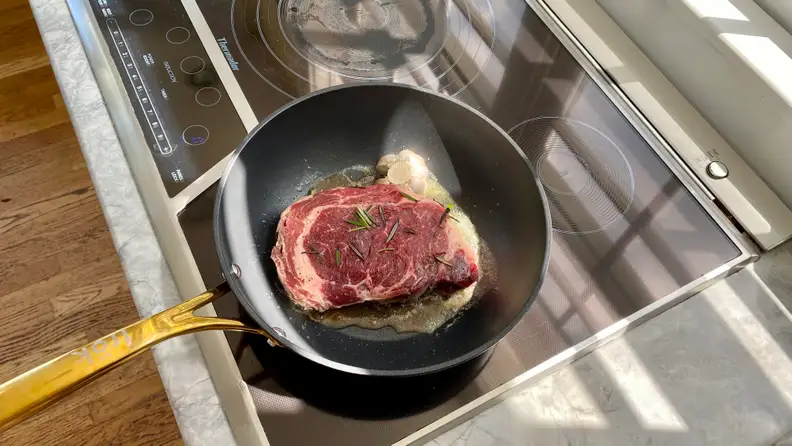 The Frök frying pan, with a piece of ribeye steak and some garlic inside, is sitting on an induction cooktop.