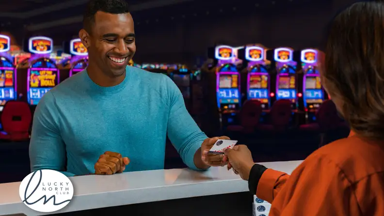 Guest presents a Lucky North Club rewards card to a casino employee at a players club desk, with colorful slot machines glowing in the background.