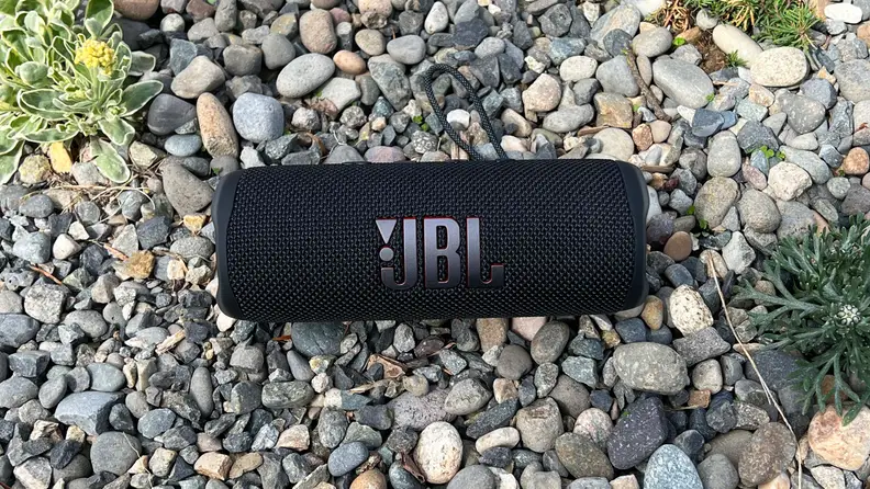 A black, cylindrical Bluetooth speaker sits in the gravel with a JBL logo showing.