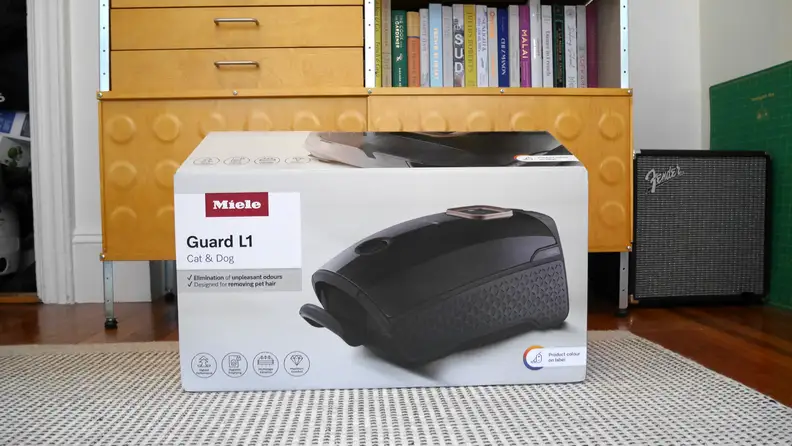 Miele Guard L1 Cat & Dog Vacuum box shown on a white and grey rug in front of a wooden cabinet with drawers and a shelf.