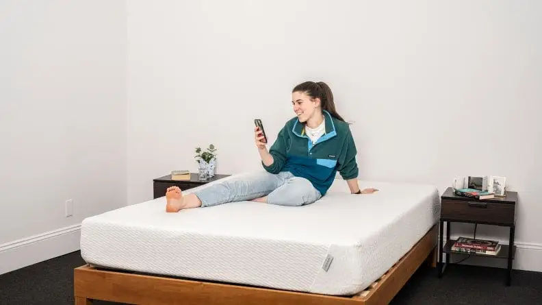 A person checks their phone while sitting on a mattress.