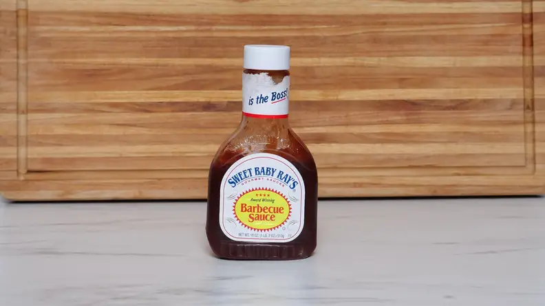 A bottle of  Sweet Baby Ray’s Barbecue  sauce next to a wood cutting board.