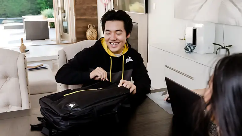A young man smiles while unzipping a Corsair backpack at a table during a casual work-from-home setup.