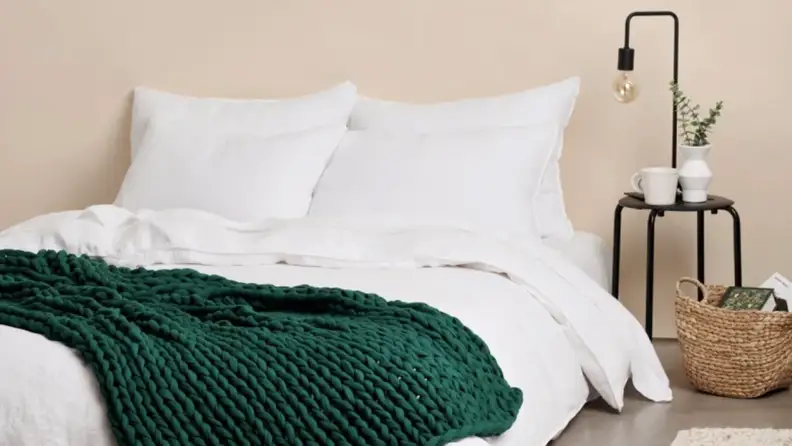 A Bearaby weighted blanket in rosemary on top of a bed, which has a white comforter and pillows.