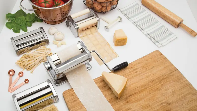 Various tools and utensils demonstrate the process of making homemade pasta. A block of cheese sits on a cutting board; tomatoes and eggs are visible at the edge of the photo.