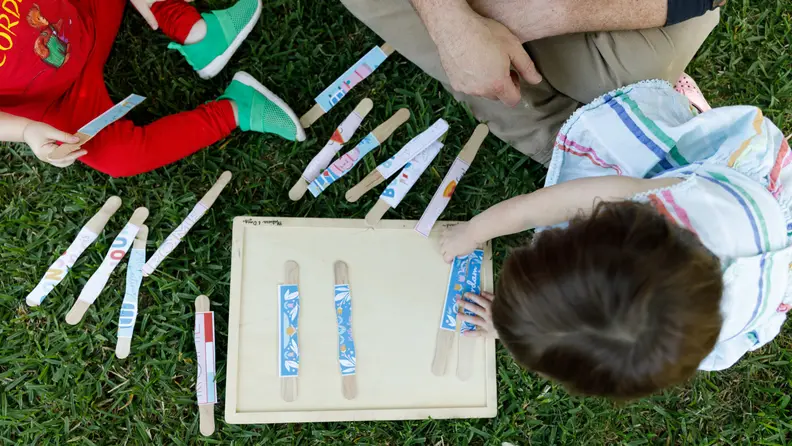 Young boy and girl playing with popsicle sticks outdoors while sitting on green grass.