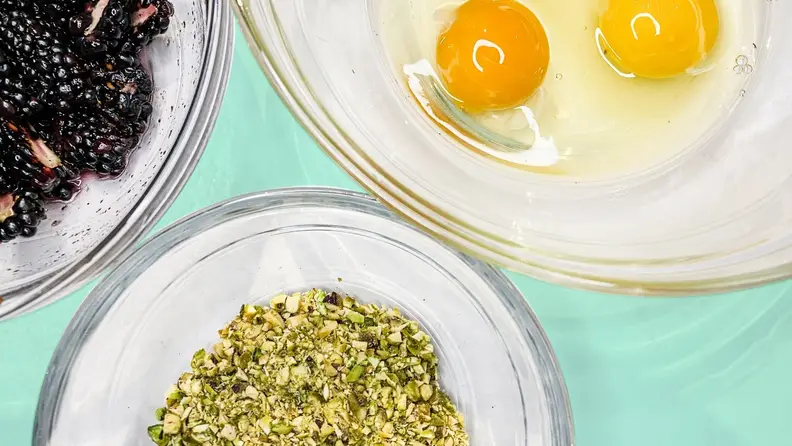 Three glass bowls filled with blackberries, crushed pistachios, and eggs.