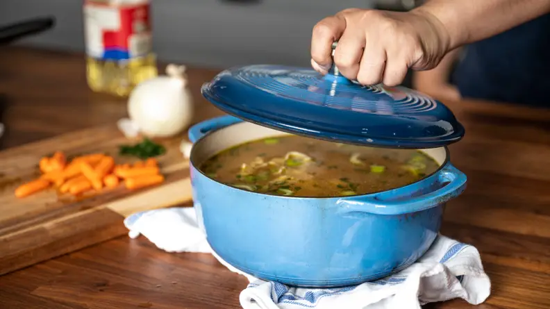 A person lifts the lid of a blue Lodge Dutch oven to reveal chicken soup inside.
