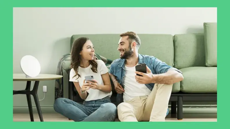 Two people sit next to each other on living room floor with light therapy lamp on nearby table.