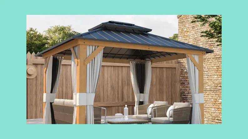 A wood gazebo with a black roof and curtains outdoors in front of a fence