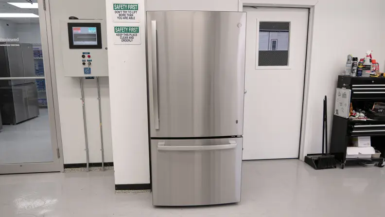 GE GDE25EYKFS Bottom-freezer Refrigerator in stainless-steel finish against white wall with safety signs and between two closed doors.