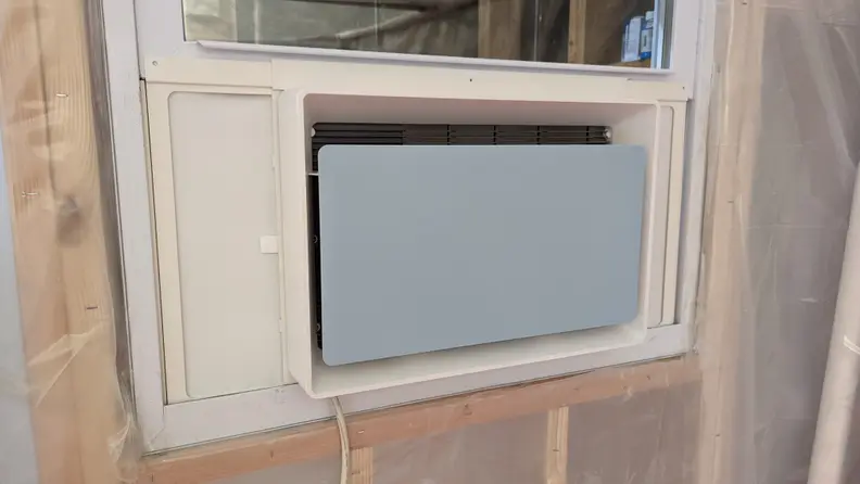 A window air conditioner with a grayish blue front panel sits in a window.