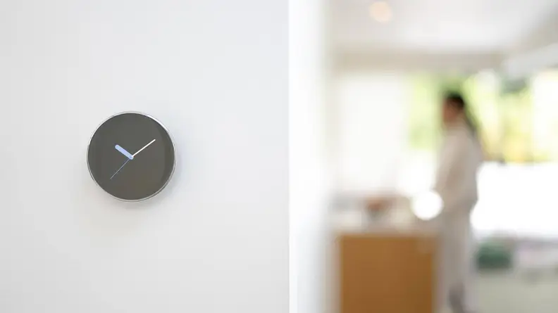The fourth-gen Nest Learning Thermostat hangs inside of a home on a white wall with the clock face displayed on the digital screen.