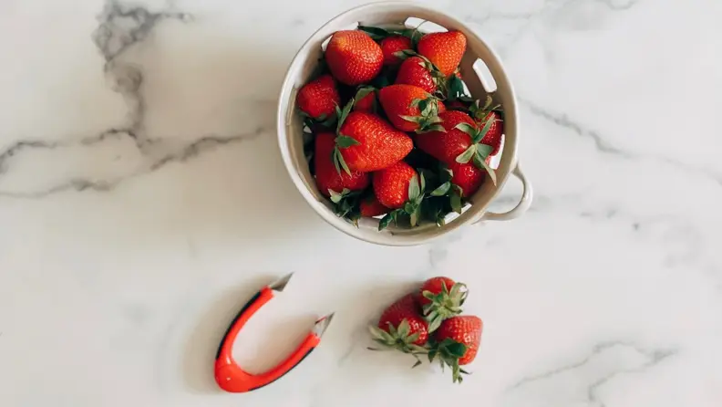 A bowl of strawberries next to the red OXO strawberry huller on a marble surface.