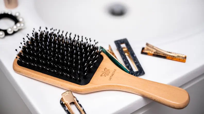 A wooden paddle brush that was named one of the best hair brushes sitting on a sink surrounded by hair clips.