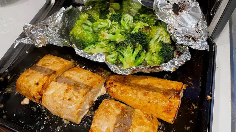 Four cooked salmon filets next to broccoli inside of aluminum foil on a pan.