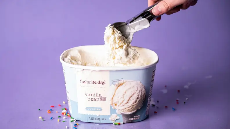 A hand scooping into a tub of vanilla ice cream on a purple background, surrounded by rainbow sprinkles.