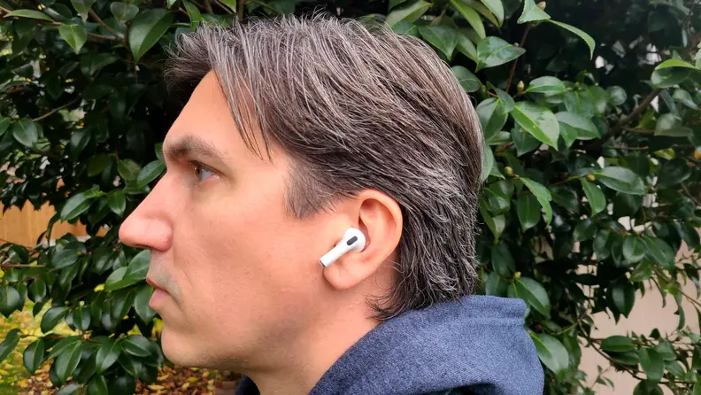 The all-white AirPod sits in the reviewer's ear with its shortened stem facing forward, set above a gret sweatshirt hood and below brown grey hair.
