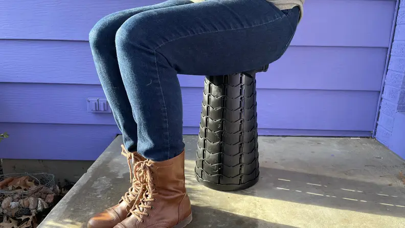 A person sitting on the Giraffe Creation Telescopic Stool outside, with a purple background.
