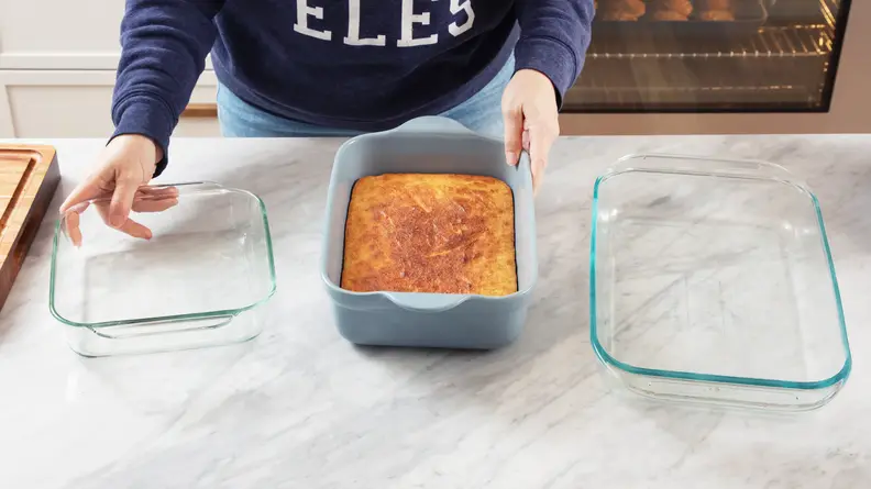 A person clutches an Our Place Bakeware pan on a gray counter with their left hand and a glass Pyrex pan with their right.