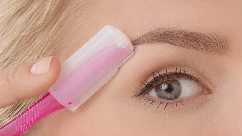 A closeup of a woman holding a dermaplane tool with a guard on it close to her eyebrow.