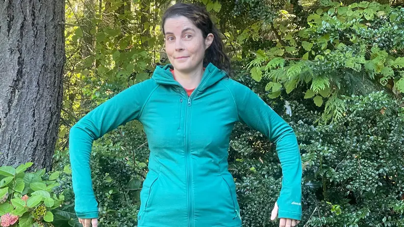 A woman outside next to trees, wearing a blue Eddie Bauer Women's High Route Full-Zip Grid Fleece Hoodie.