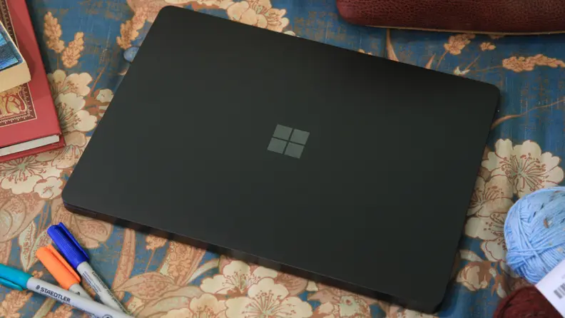 An overhead view of a closed Microsoft Surface Laptop 7th Edition showing its keyboard on a flower-patterned cloth surface surrounded by markers, books, and yarn.