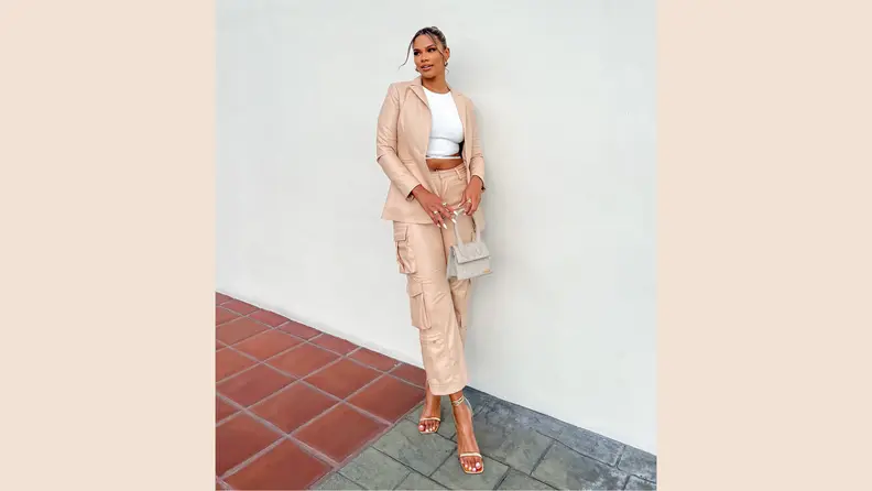 A woman wearing a beige leather suit with cargo pants, a white shirt, and beige sandals. She holds a small handbag.