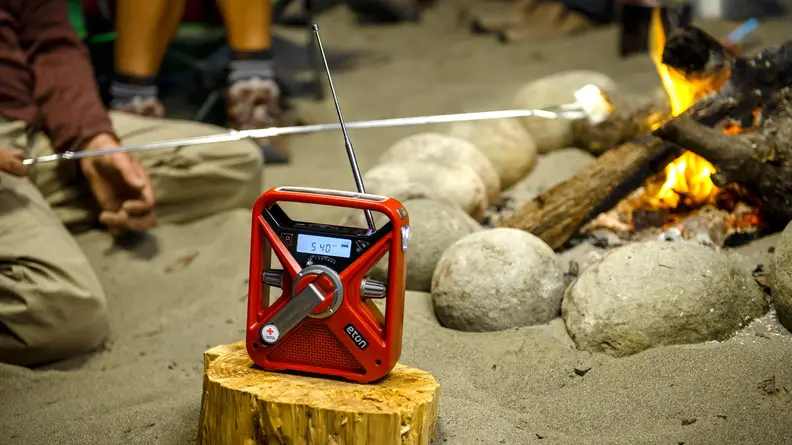 Red Etón FRX3+ Multi-Powered Weather Alert Radio sitting on top of wooden log next to person roasting marshmallow on campfire.