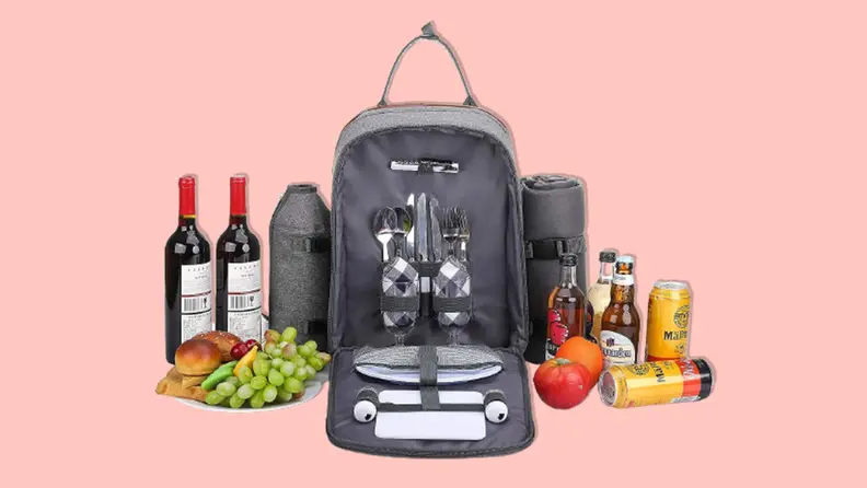 An Allcamp Outdoor Gear Picnic Backpack filled with plates and silverware in the middle of two bottles of wine, fruits, a several soft drinks on a pink background.