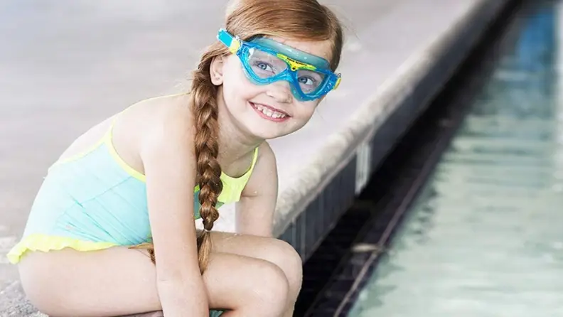 A girl sits on the side of a pool smiling with her kids swim goggles on