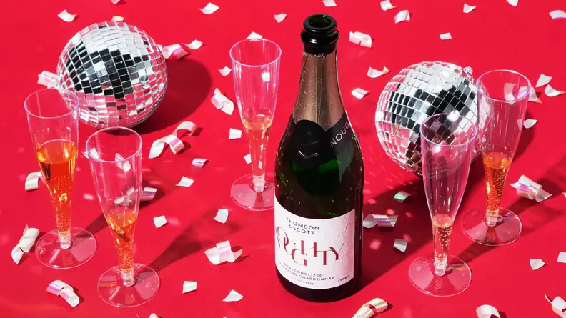 A bottle of Noughty Alcohol-Free Sparkling Chardonnay on a red background with confetti and disco balls.