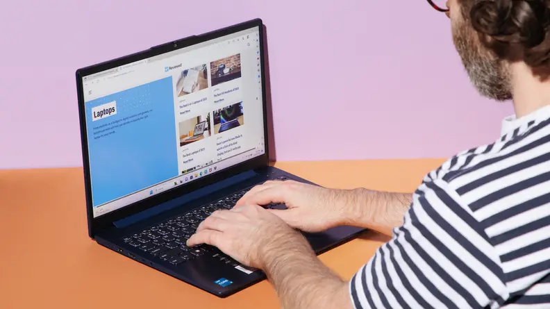 A person using an open and powered on laptop against a pastel purple background.