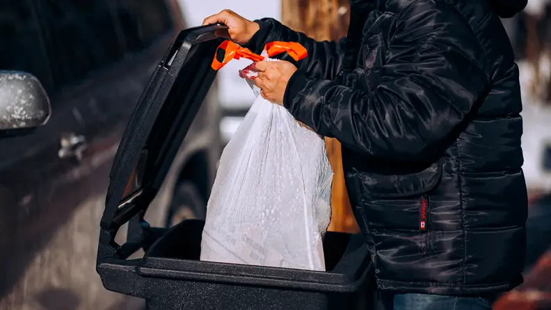 A person putting a white trash bag in a black outdoor trash can.