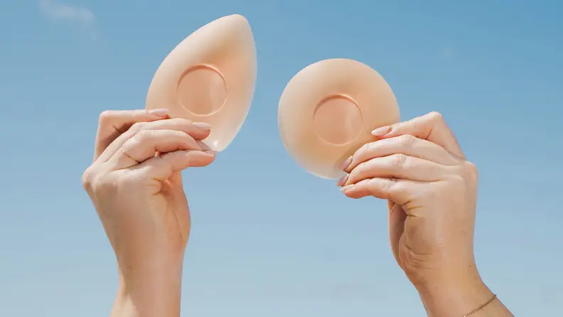 Hands holding two different shapes of Cakes nipple covers against a clear blue sky.