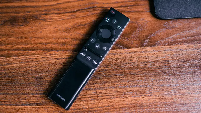A black, minimalist Samsung remote control sits on a table rich with wood grain