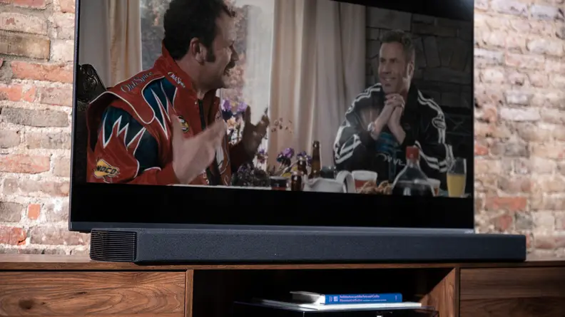 The black Q950a sits on a woodgrain cabinet in front of a large TV, with brick background.